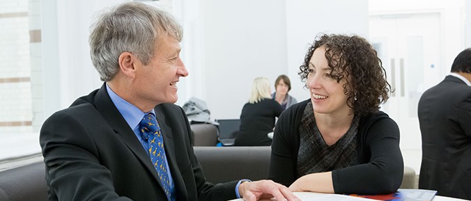 Two seated colleagues wearing suits have a conversion in a large room. 