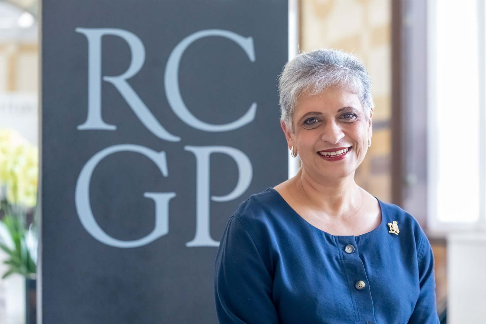 Professor Kamila Hawthorne standing by a large navy blue RCGP sign wearing a blue top.