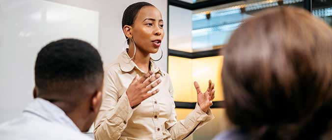 A lady giving a talk to a room of people, a man and woman facing her, engaging.