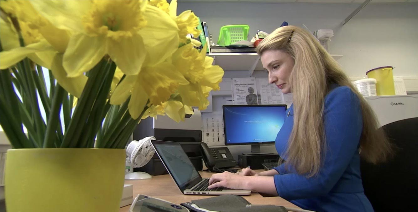 An image of Dr Catherine Millington-Sanders working at her desk with a bunch of yellow daffodils in the foreground.