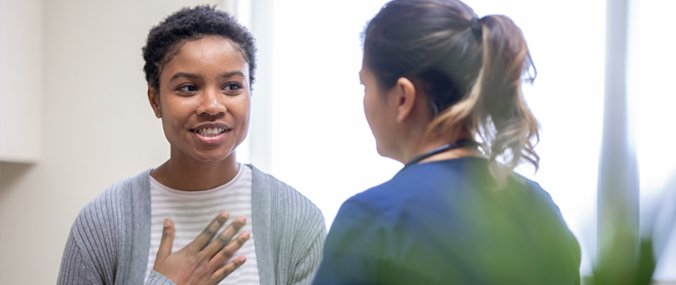 A woman with short hair converses with a health professional in a white clinic, holding one hand to her chest. 