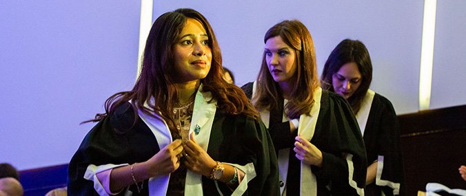 Three women wearing dark graduate robes during a ceremony.