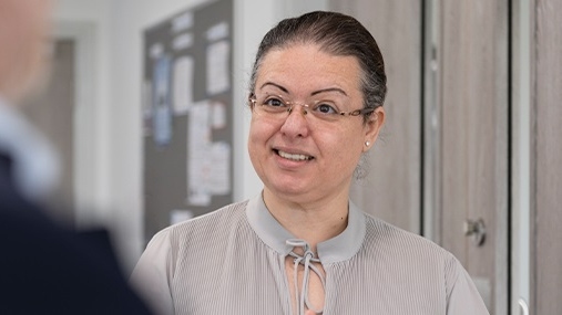 A woman wearing glasses and a grey blouse smiles at a person in the distance. 